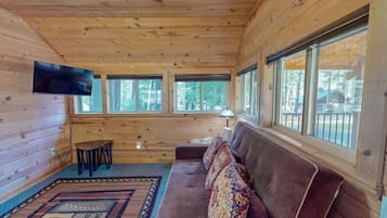 A cabin interior with a wooden ceiling, a sofa, a TV, and large windows.