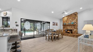 Eine moderne Küche mit einer großen Kochinsel, einem Essbereich mit Holzstühlen, einem Kamin aus Stein und Blick auf einen Swimmingpool.