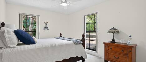 Un dormitorio con cama, mesita de noche, ventilador de techo y ventanas amplias.