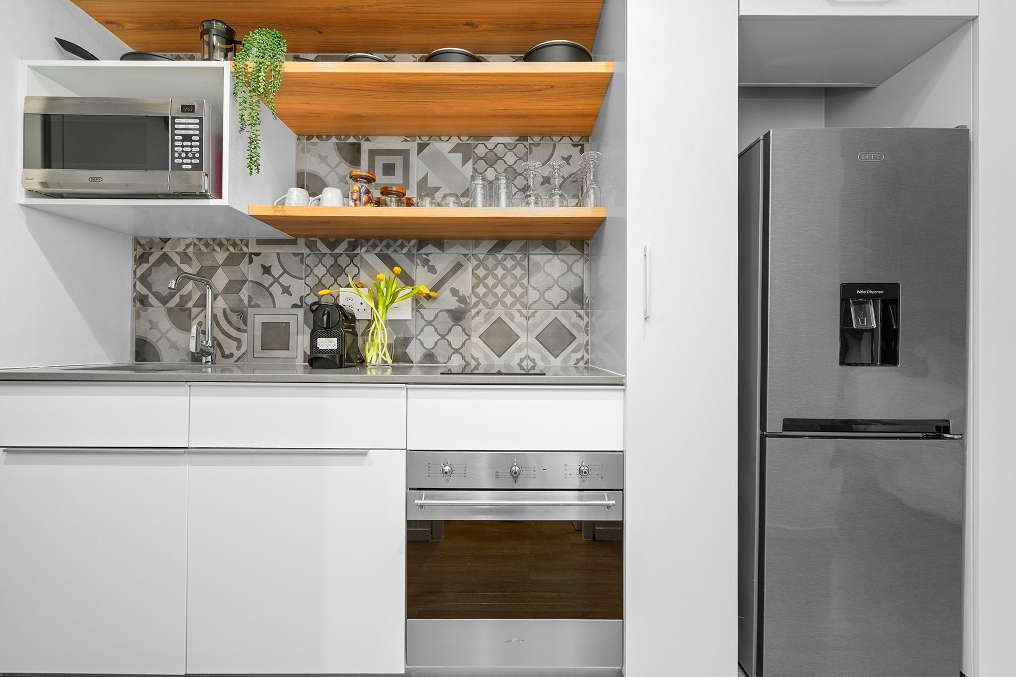 A modern kitchen with a stainless steel refrigerator, microwave, and oven. There is a wooden shelf above the microwave and a tiled backsplash.