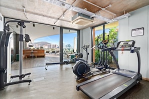 Ein Fitnessstudio mit Laufbändern, Crosstrainern und Krafttrainingsgeräten. Es gibt große Fenster mit Blick auf eine Stadtlandschaft und ein Gebirge.