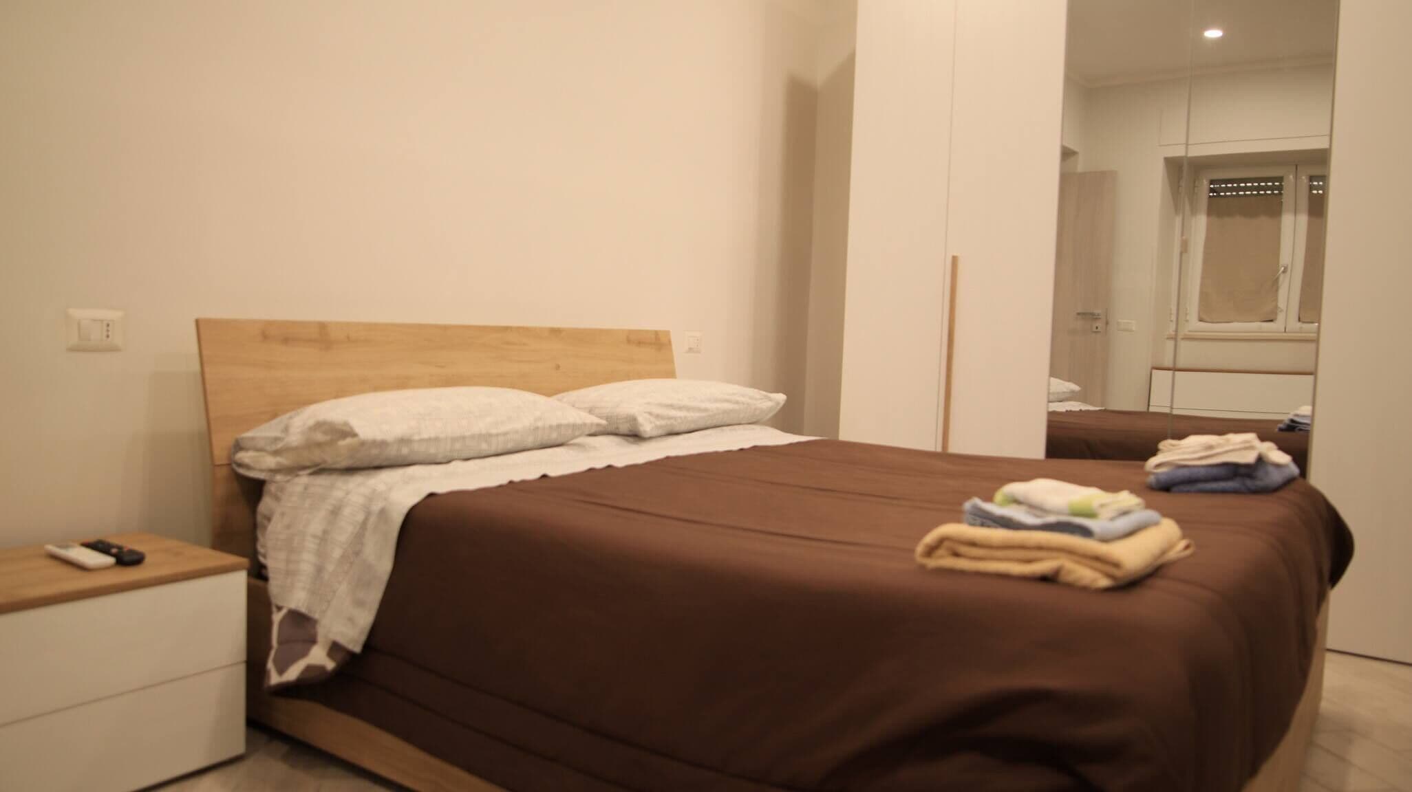 A neatly made bed with a brown bedspread and white pillows, a bedside table with a remote control, and a mirror reflecting the room.