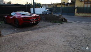 Une voiture de sport rouge est garée sur un terrain recouvert de gravier, avec un mur de béton et une clôture en arrière-plan.