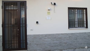 Une façade d’immeuble avec une grande porte en bois, une fenêtre avec des barreaux en métal et un système d’interphone fixé au mur.