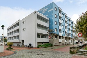 Un immeuble d’appartements moderne aux façades aux couleurs variées, allant du blanc au bleu, situé sur une rue pavée, avec un vélo garé devant.