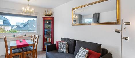 Un salon avec un canapé foncé, une bibliothèque rouge, une table à manger avec des chaises et un grand miroir au mur.
