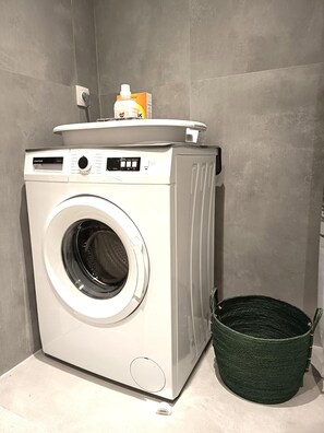 Un lave-linge compact et moderne, doté d’un panneau de commande, d’un compartiment à lessive et d’un panier vert placé à côté.