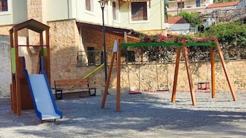 Um parquinho com escorrega e balanços, com um muro de pedra e edifícios ao fundo.