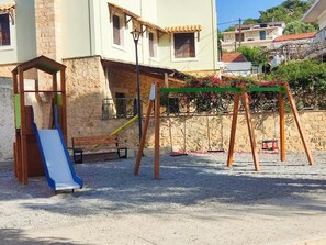 A playground with a slide and swings, set against a stone wall and buildings.