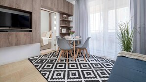 Een moderne woonkamer met een aan de muur bevestigde televisie, een eethoek met stoelen en een tafel, en een groot raam met gordijnen.