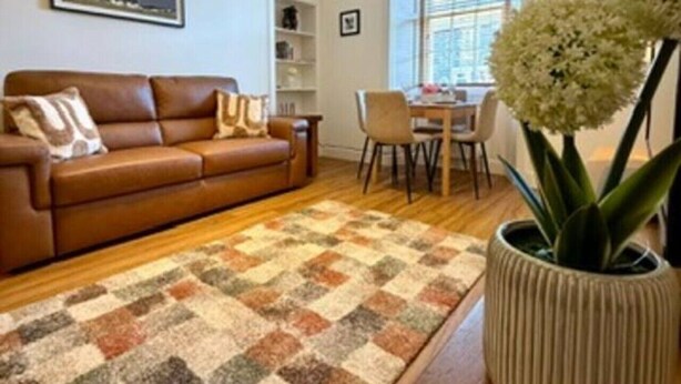 A living room with a brown leather sofa, a patterned rug, a dining table with chairs, and a potted plant. - Elliot Cottage Your Retreat in Heart of Peebles (Scottish Borders)