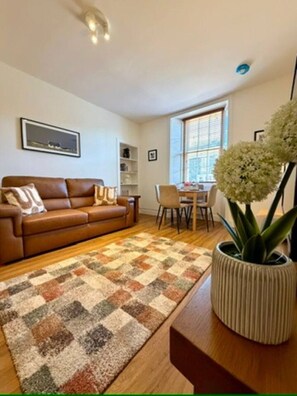 A living room with a brown leather sofa, a patterned rug, a dining table with chairs, and a potted plant. - Elliot Cottage Your Retreat in Heart of Peebles (Scottish Borders)