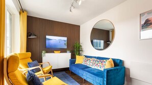 A modern living room with a blue sofa, yellow armchairs, a flat-screen TV, a round mirror, and a wooden wall panel. - Rigg & Rest - Central Bowness, Balcony & Parking (Bowness-on-Windermere)