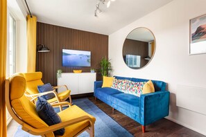 Ein modernes Wohnzimmer mit einem blauen Sofa, gelben Sesseln, einem Flachbildfernseher, einem runden Spiegel und einer Holzvertäfelung.