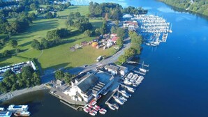 An aerial view of a marina with boats docked, a large building with a flat roof, and surrounding greenery. - Rigg & Rest - Central Bowness, Balcony & Parking (Bowness-on-Windermere)