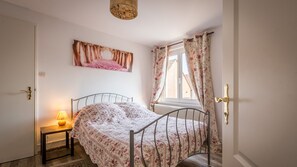 Ein Schlafzimmer mit Bett, Nachttisch mit Lampe, einem Fenster mit Vorhängen und einem an der Wand hängenden Kunstwerk.