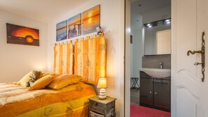 Un dormitorio con cama, mesita de noche con lámpara, una pared con obras de arte y un baño visible a través de una puerta abierta.
