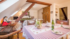 Un comedor con mantel a cuadros, sillas de madera, un aparador rústico y un espacio elevado con una muñeca y un cofre de madera.