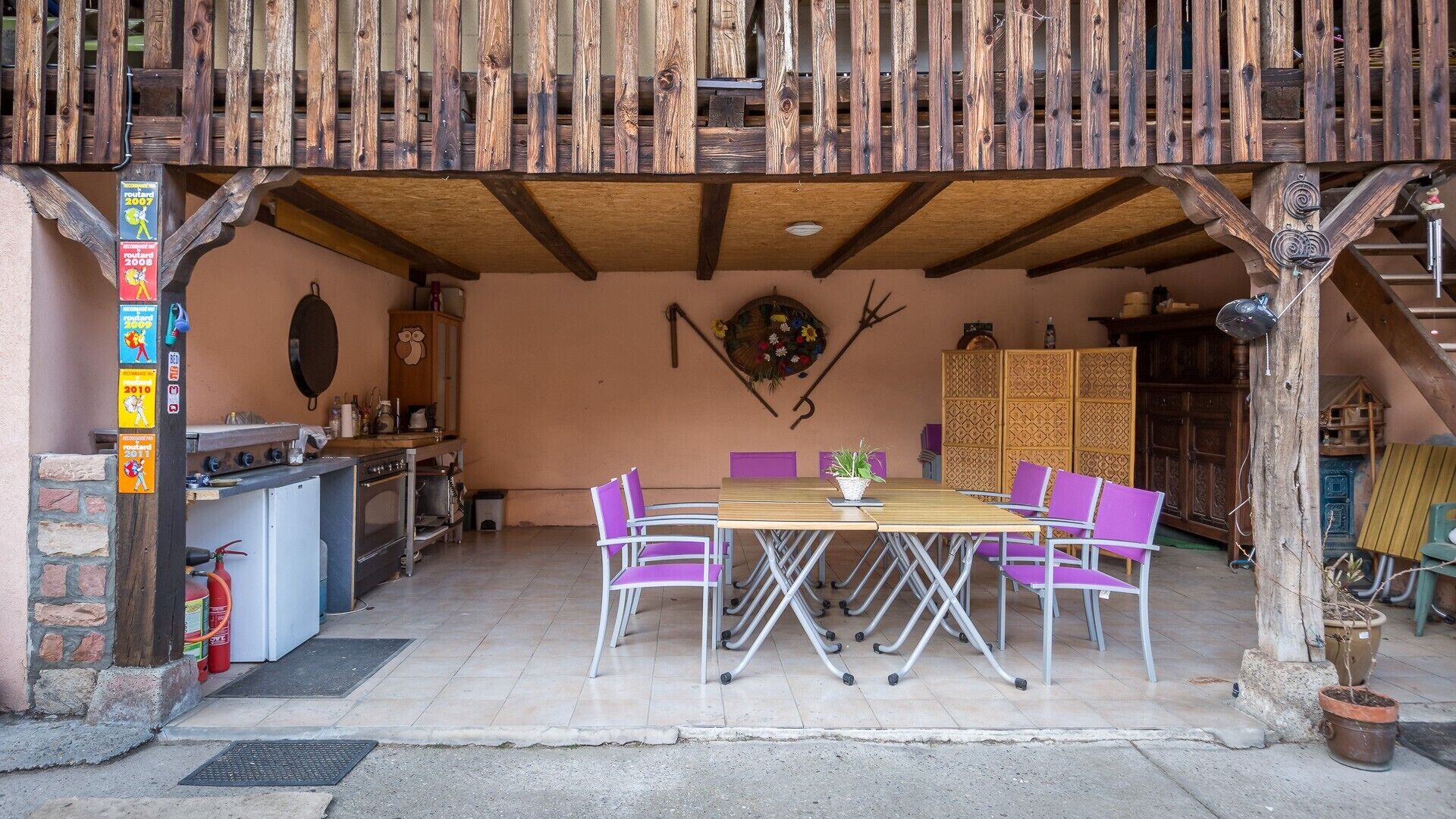 Une terrasse extérieure avec une table en bois et des chaises violettes, un coin cuisine équipé d’une cuisinière et de placards, ainsi qu’un élément décoratif mural.
