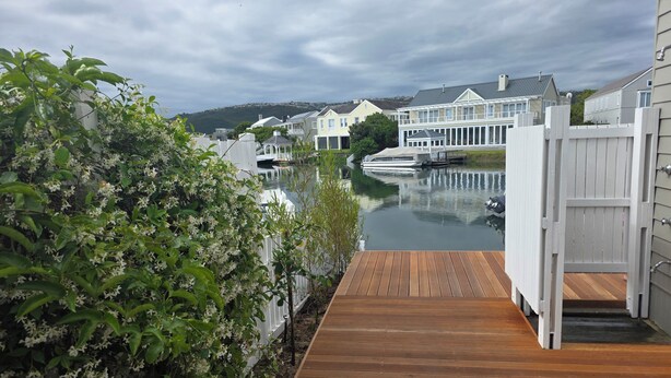 A wooden deck leading to a gated area with a view of a calm water body and houses in the background.