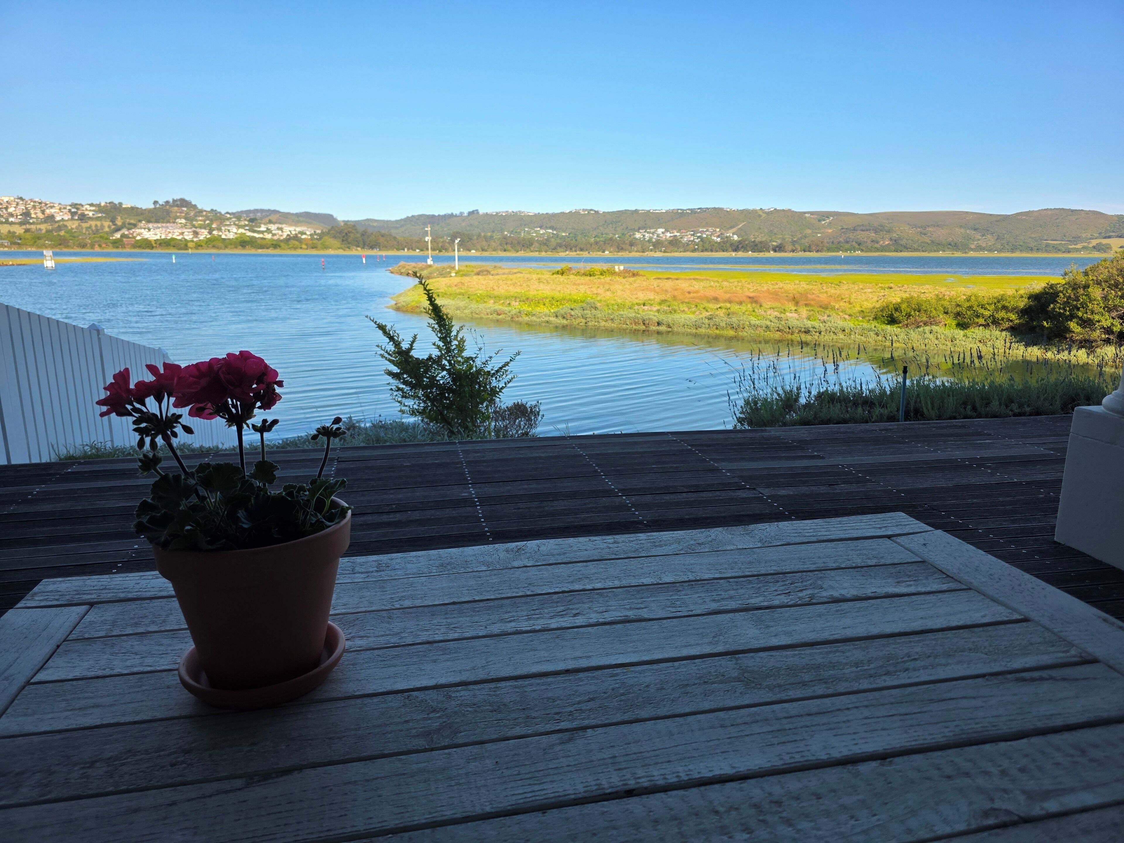 Een houten terras met een bloempot en uitzicht op een meer met boten en heuvels in de verte.