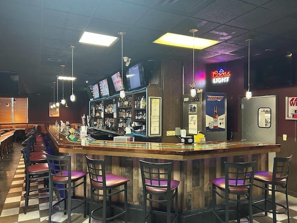 Een bar met een toonbank, barstoelen en een neonbord.