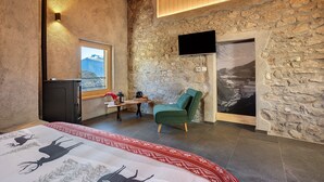 A room with a stone wall, a mounted TV, a green chair, a bed with a patterned bedspread, and a window with a mountain view. - Chalet 'Al Volt' with Mountain View, Private Terrace and Garden (Voiandes)