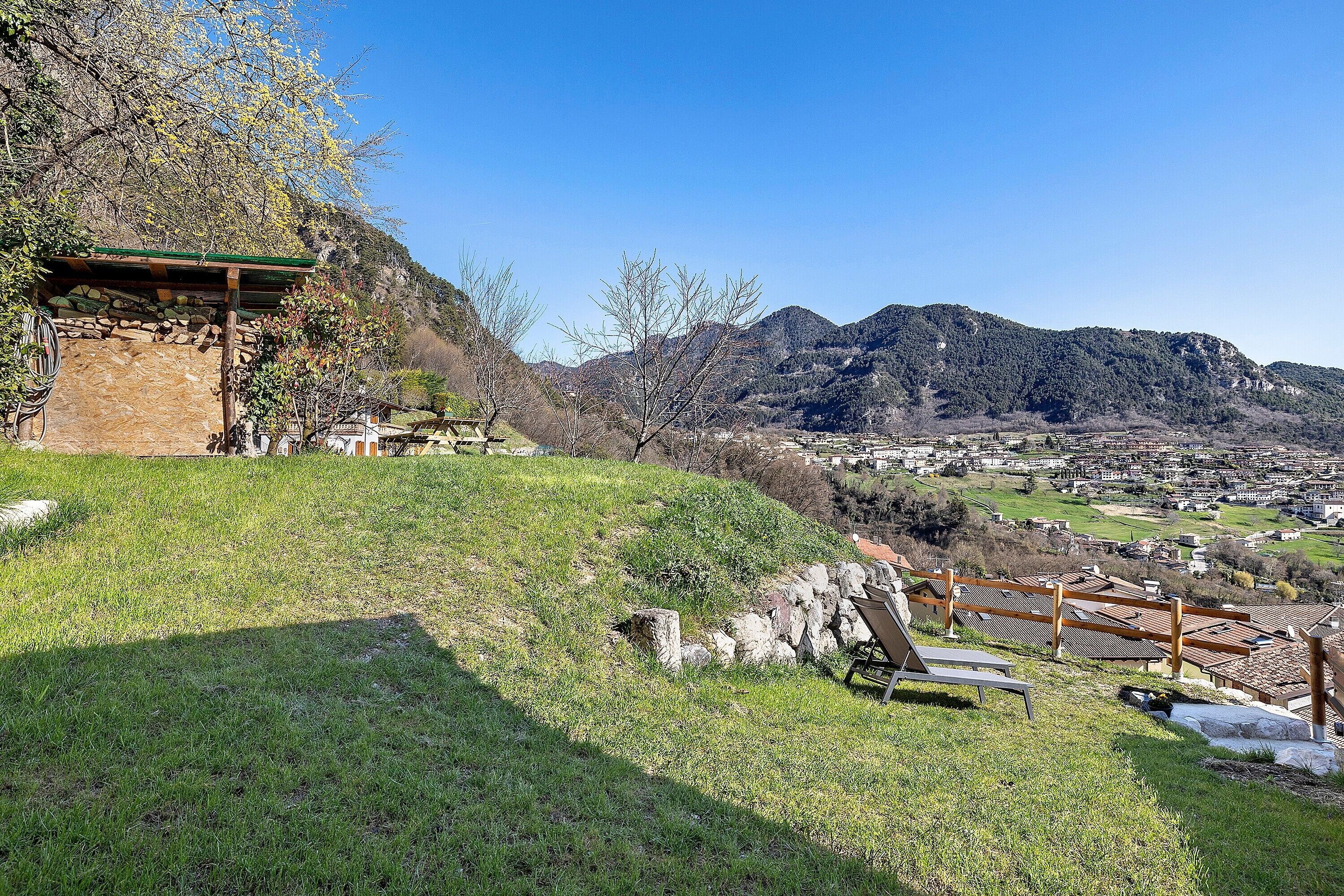 Una colina cubierta de hierba con una estructura de piedra, bancos de madera y vistas a un pueblo y montañas al fondo.