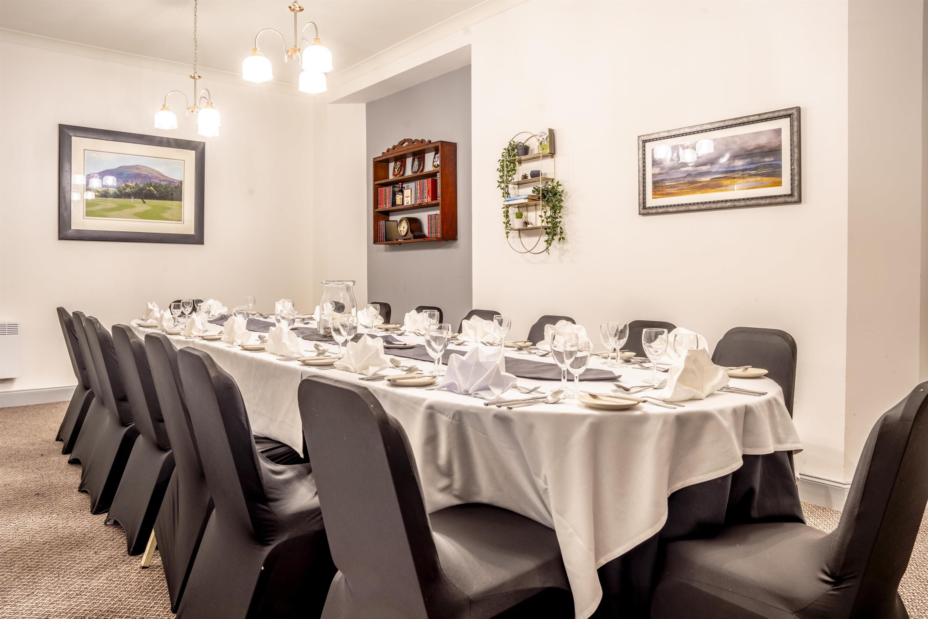 A conference room with a long table set for a meeting, surrounded by chairs, with a bookshelf and framed pictures on the walls.