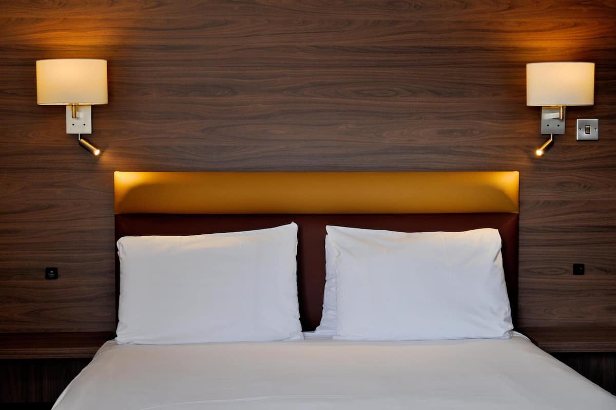 Habitación de hotel con cabecera de madera, dos almohadas blancas y lámparas fijadas a la pared.