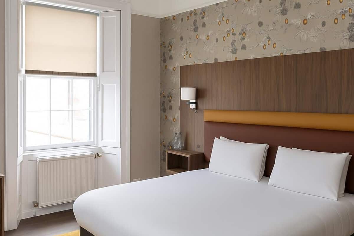 Habitación de hotel con una cama grande, una ventana con persianas, un cabecero de madera, una mesita de noche con lámpara y papel tapiz estampado.
