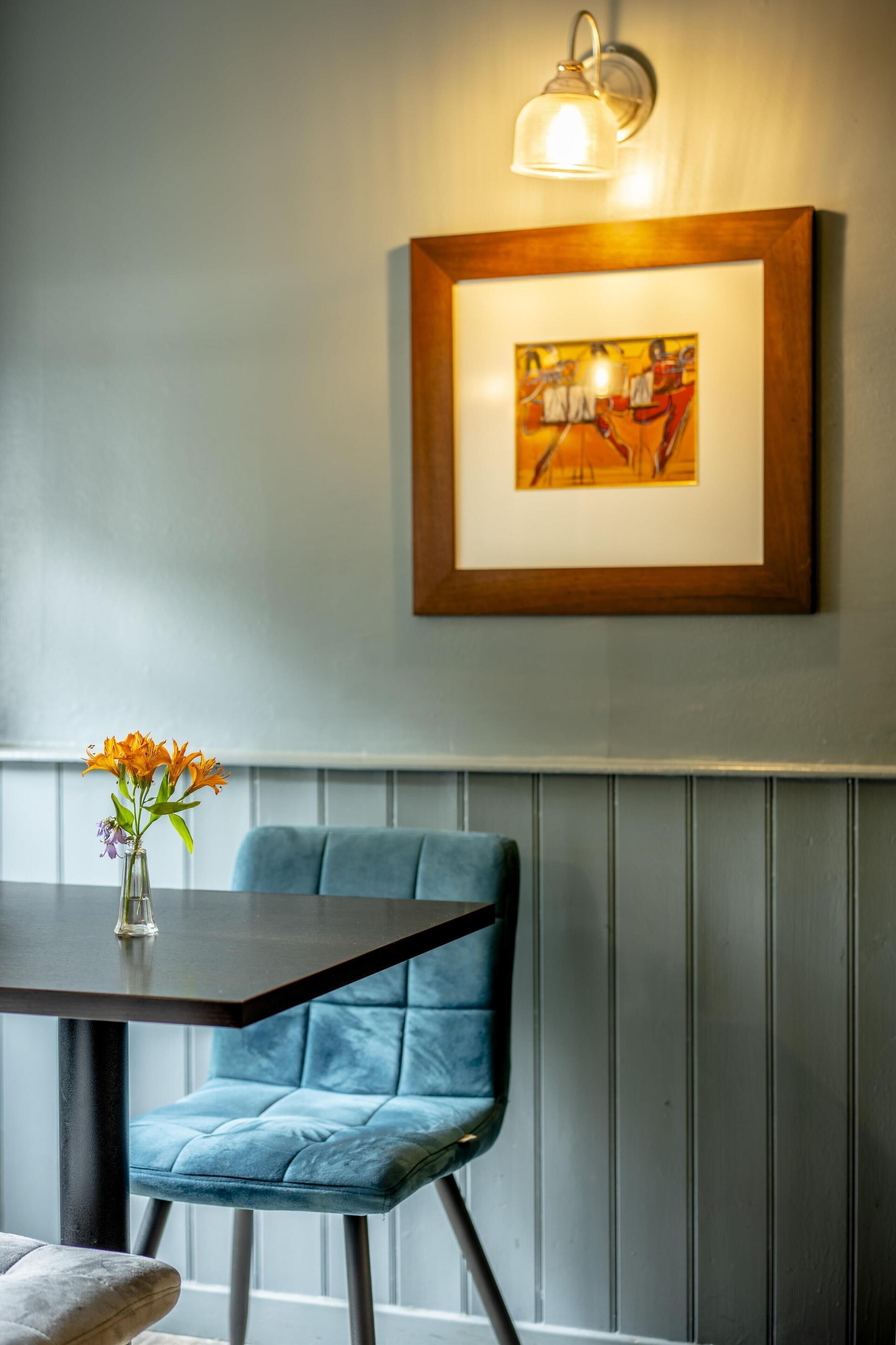 A dining area with a table, a blue upholstered chair, a vase with flowers, and a framed artwork on the wall.
