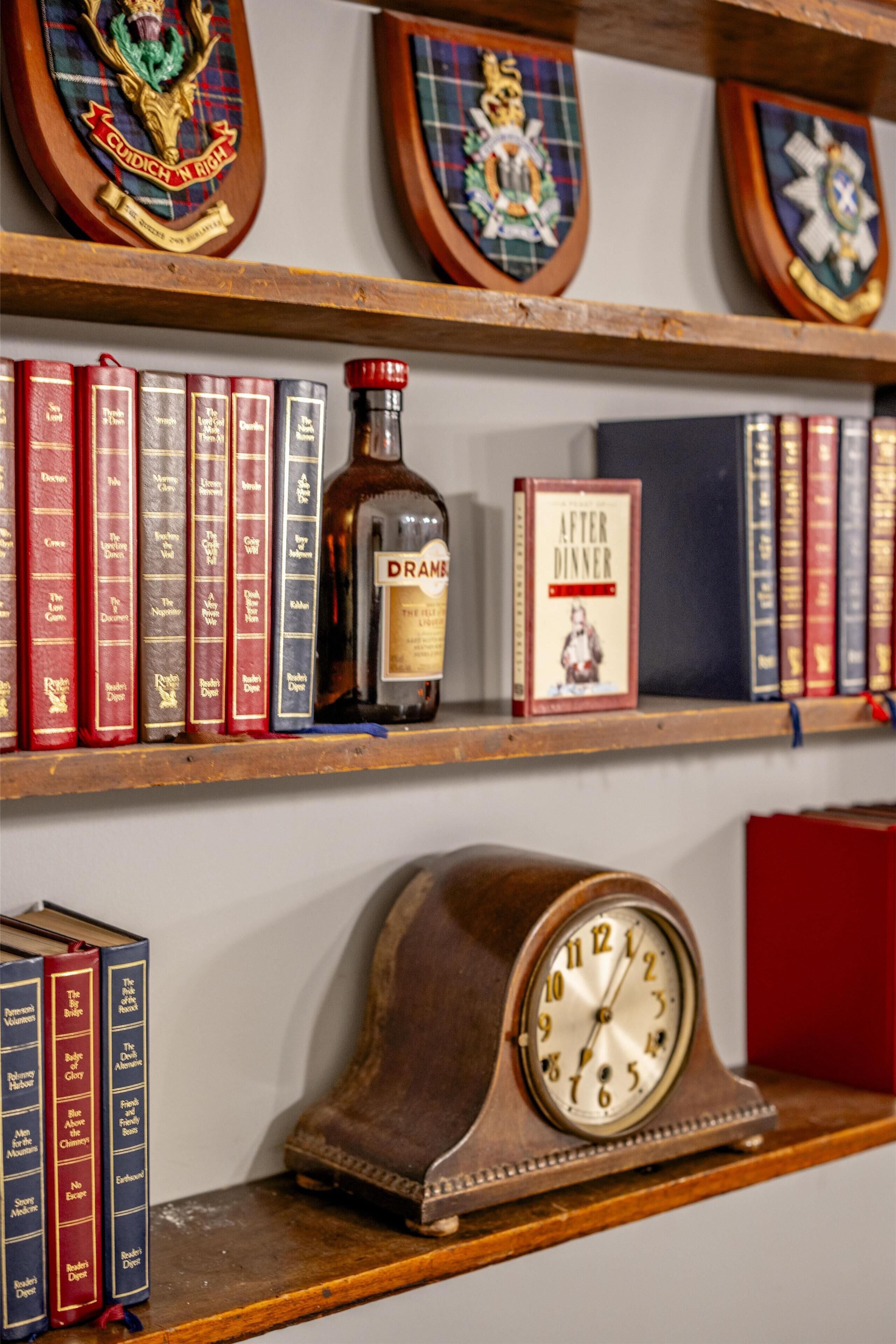 A bookshelf with books, a bottle of Drambuie, a clock, and framed coats of arms.