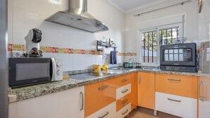 A kitchen with a microwave, stove, and oven, white and orange cabinets, a stainless steel range hood, and a window with a view of greenery outside. - Vacation Apartment in Chiclana with Pool and Garden (Chiclana de la Frontera)