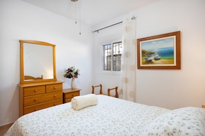 A bedroom with a bed, a dresser, a mirror, a painting, and a window with a view. - Vacation Apartment in Chiclana with Pool and Garden (Chiclana de la Frontera)