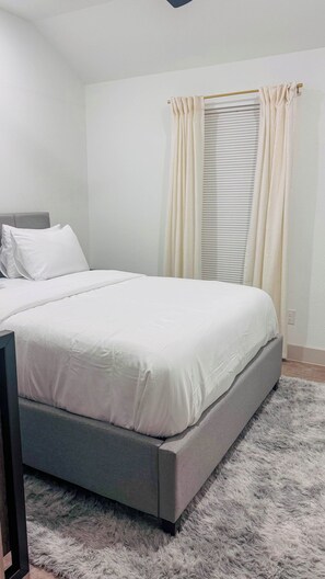 A bedroom with a bed, a nightstand, a window with curtains, and a fluffy rug. - Little Nyssa Home Away From Home (Houston)