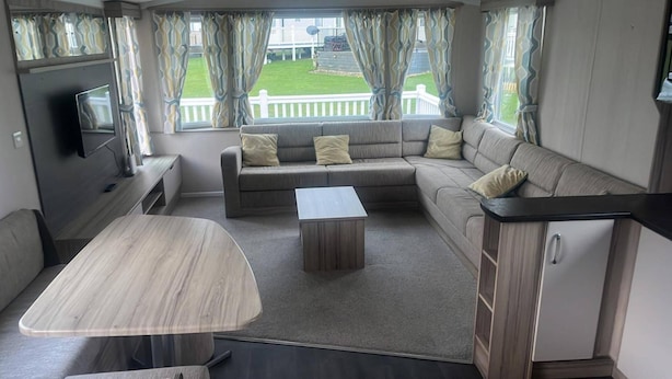 A cozy RV interior with a sofa, a small table, a TV, and a view of a grassy yard through the windows. - Caravan- Sleeps 6, 2 Bedrooms - Thurston Manor (Dunbar)