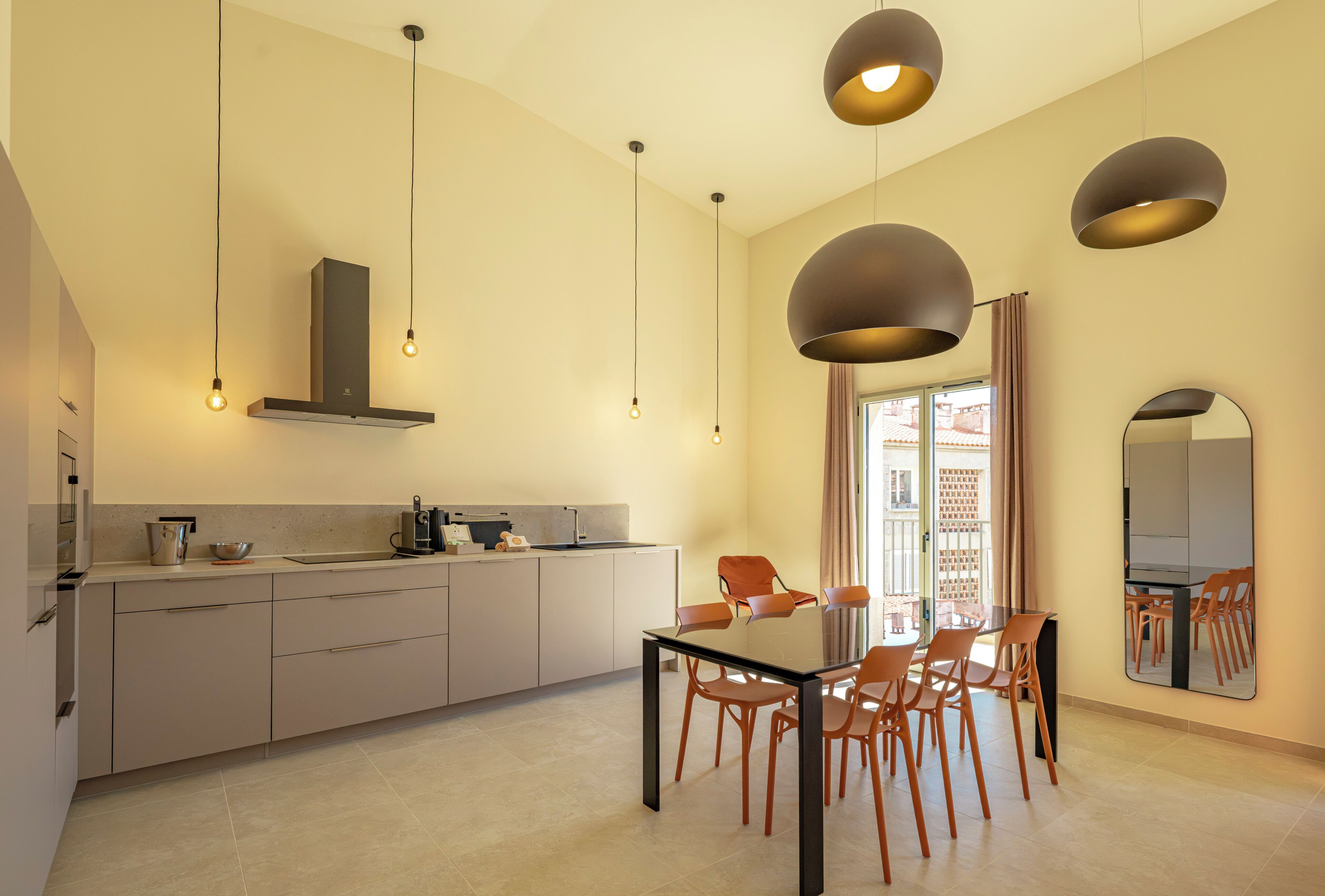 A modern kitchen with a dining area, featuring a central island, pendant lighting, and a large window with curtains.