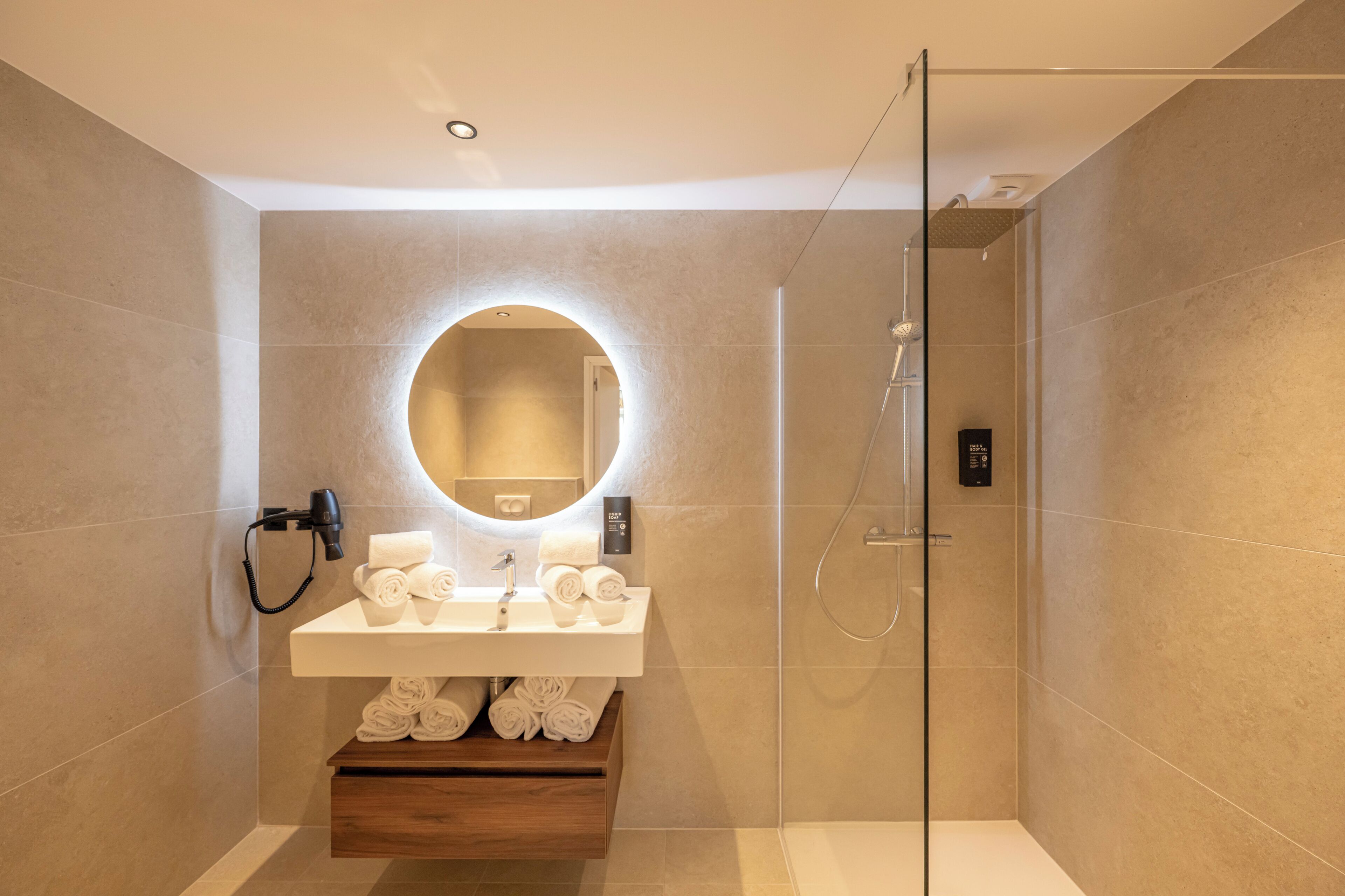 Een moderne badkamer met een douchecabine van glas, een ronde spiegel en een houten wastafelmeubel met netjes opgerolde handdoeken.