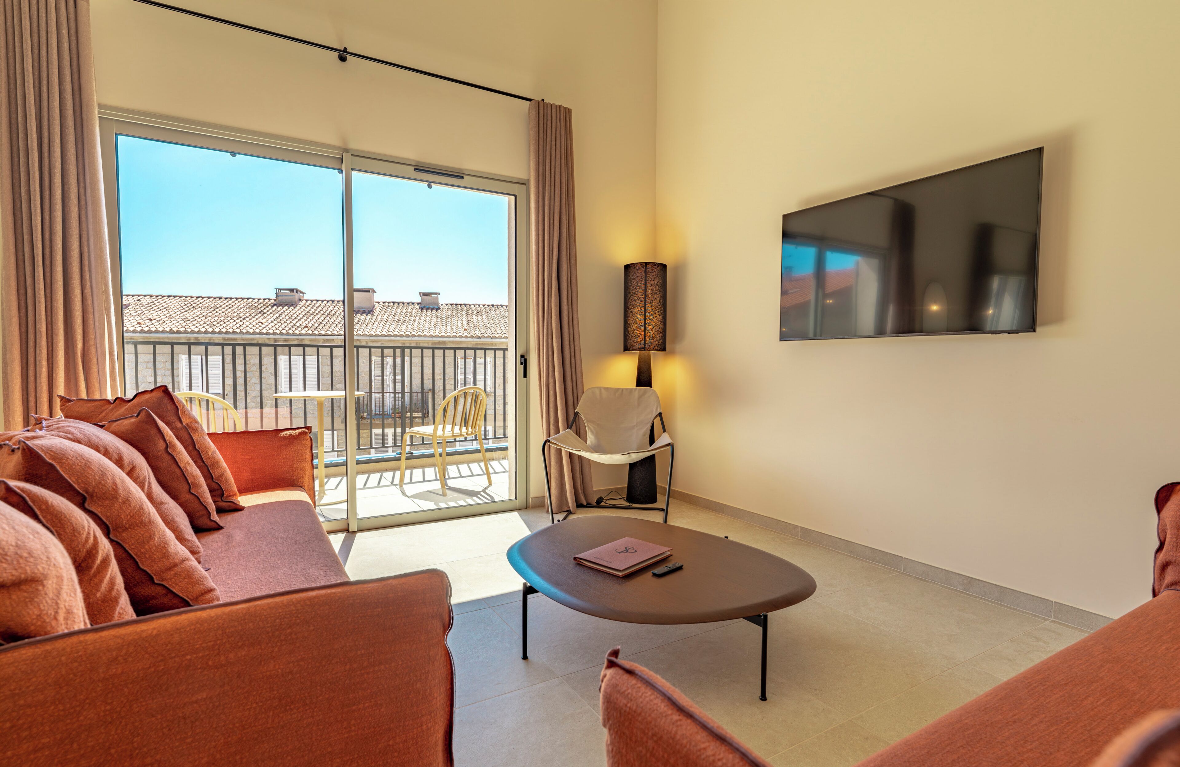 Ein Hotelzimmer mit einer Couch, einem Tisch, einem Stuhl, einem Fernseher und einem Balkon mit Blick auf Gebäude.
