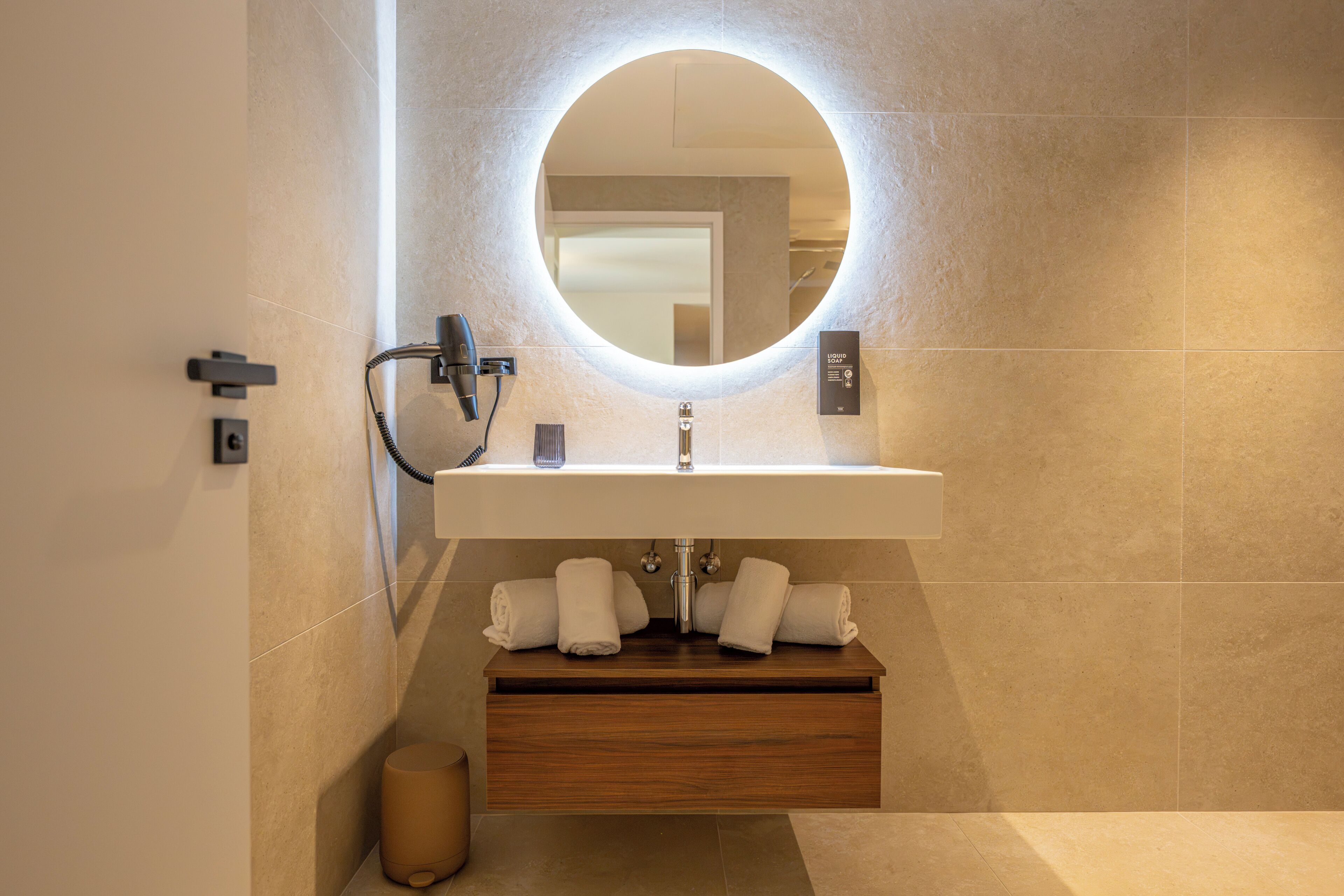 A modern bathroom with a round mirror, a white sink, and a wooden vanity.
