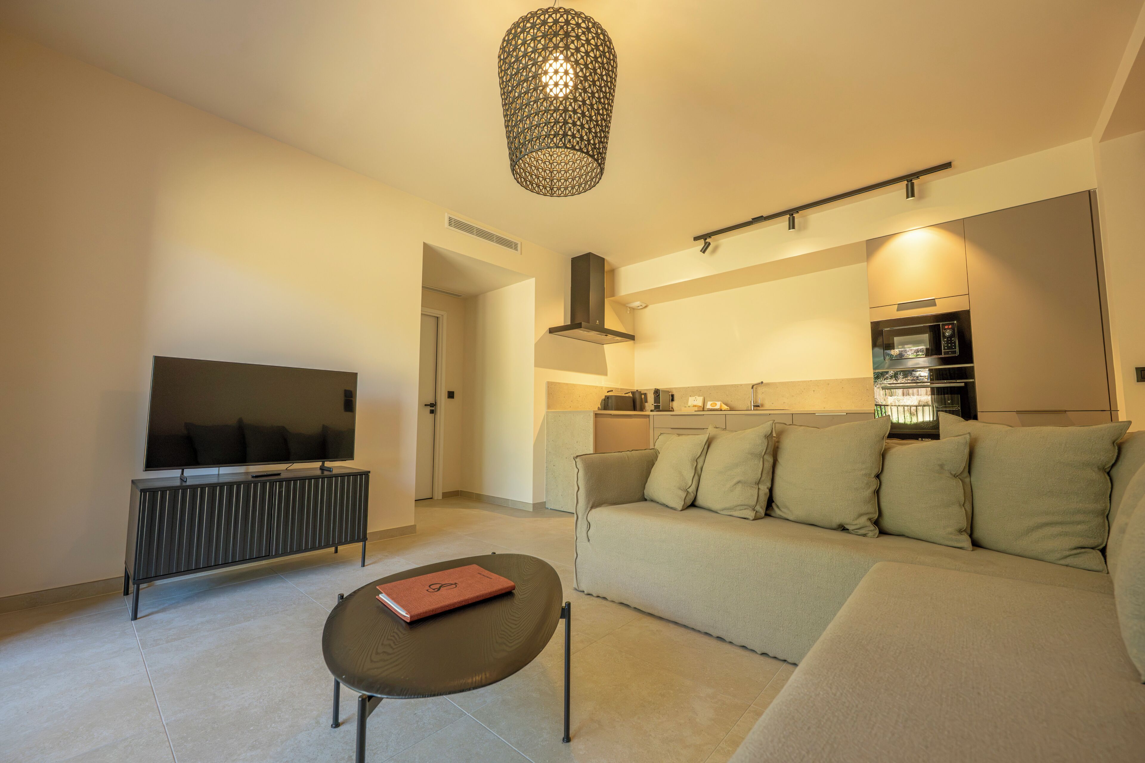 Ein modernes Wohnzimmer mit Flachbildfernseher, einer Couch, einem Couchtisch und einer Küche im Hintergrund.