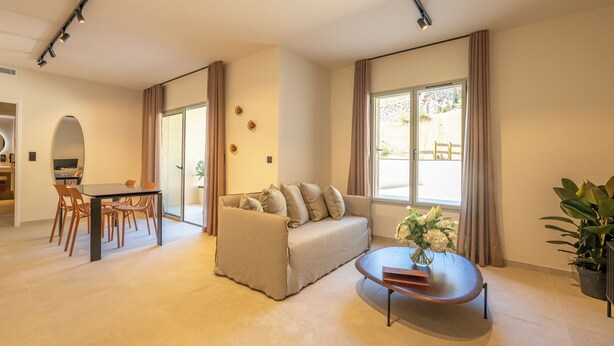 Una sala de estar moderna con sofá, mesa de comedor y mesa de centro. Hay un ventanal con cortinas, un espejo y una planta en maceta.
