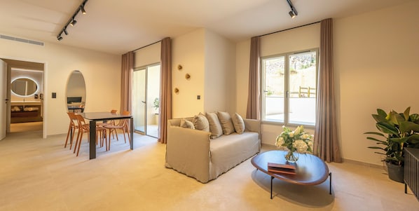 A modern living room with a sofa, a dining table, and a coffee table. There is a large window with curtains, a mirror, and a potted plant.