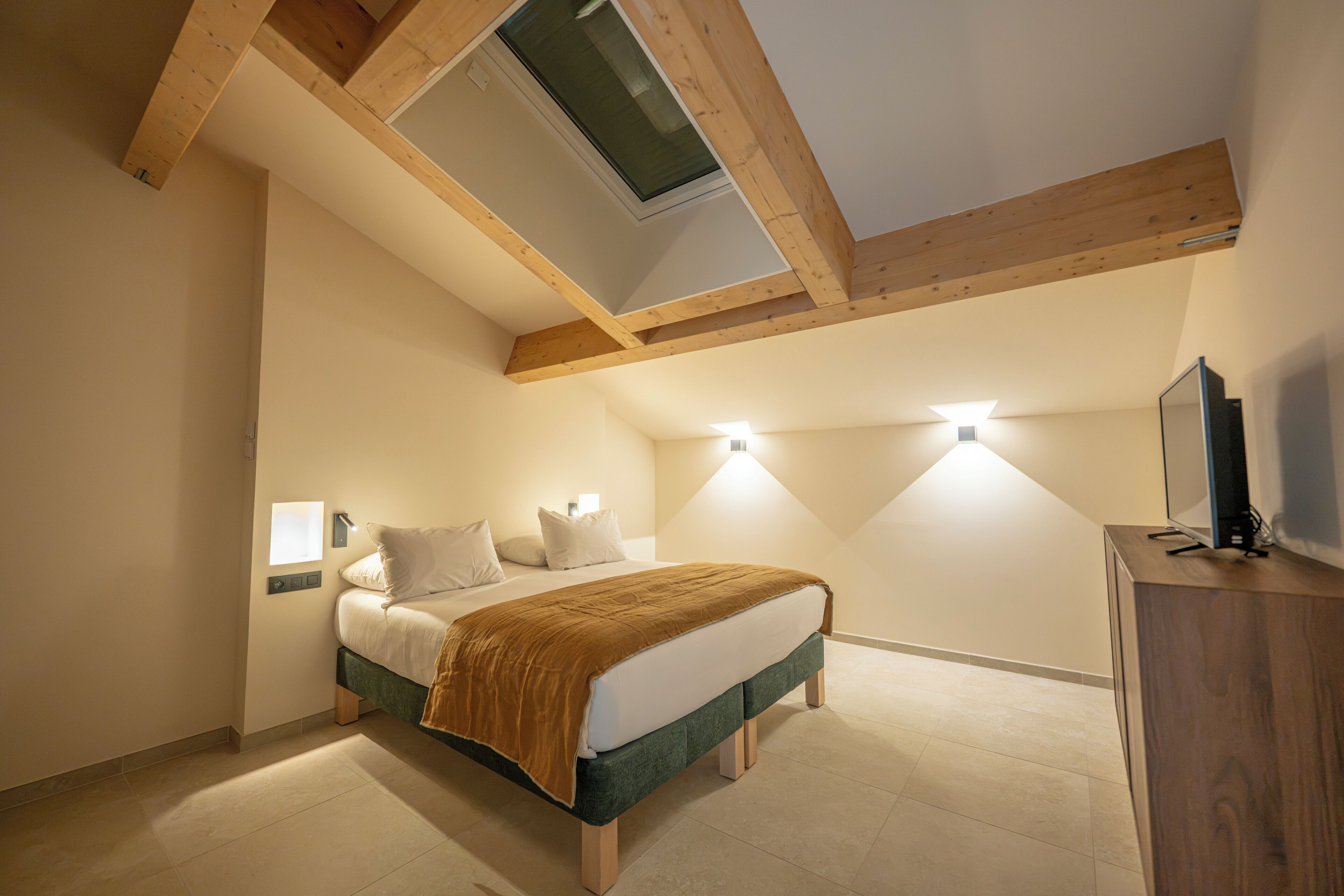 A bedroom with a sloped ceiling, a bed with a green frame, a wooden desk, and a television on the desk.