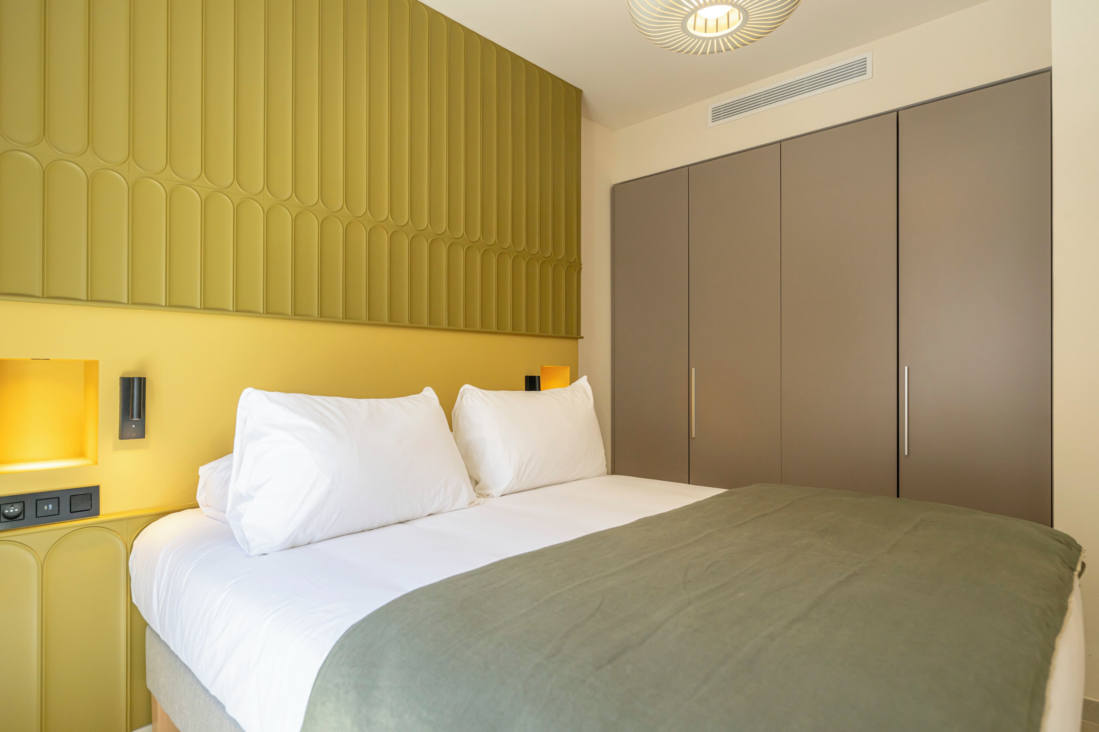 A hotel room with a bed, bedside tables, a wall-mounted light, and a wardrobe.
