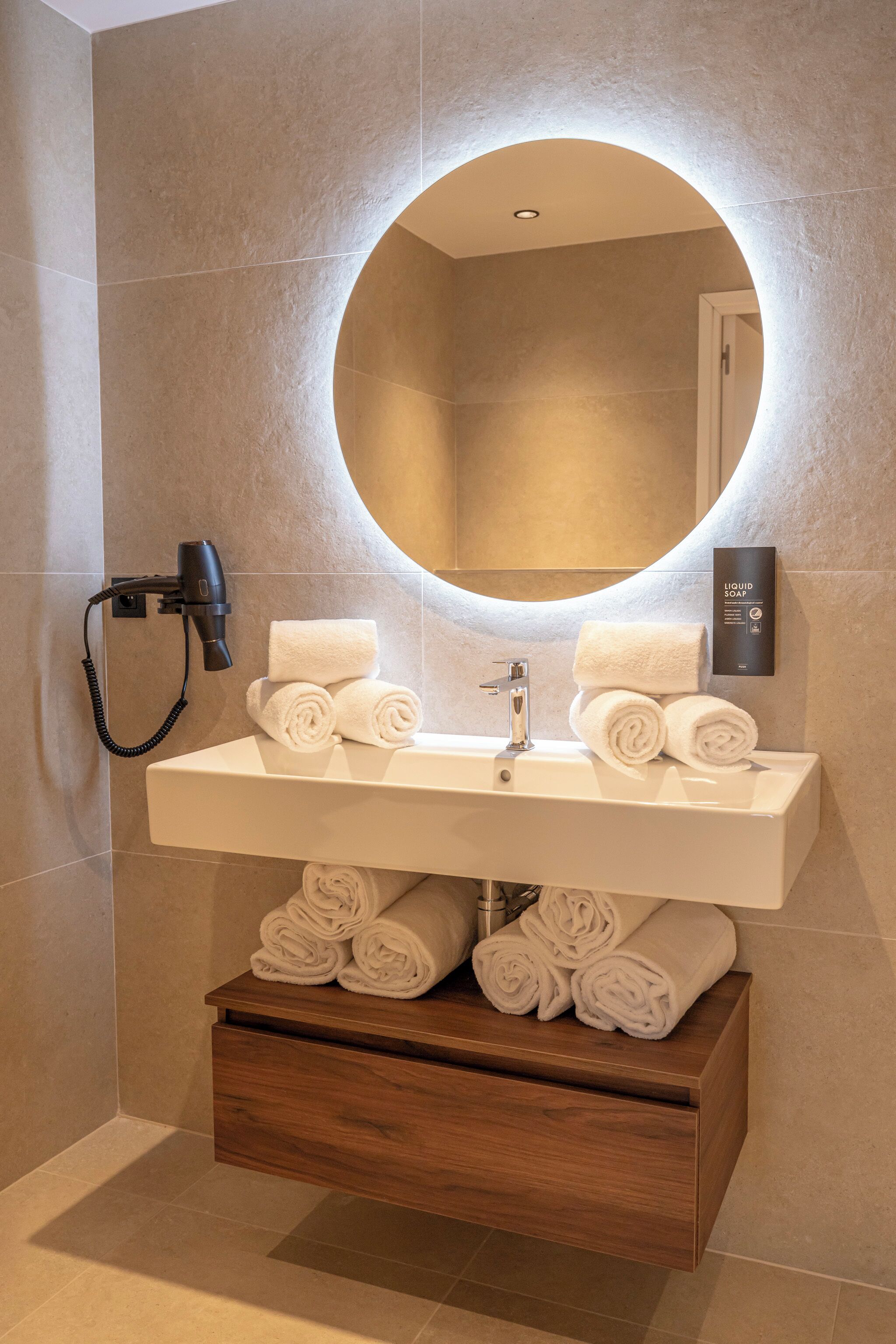 A bathroom with a round mirror, a sink, and a wooden storage unit with towels.