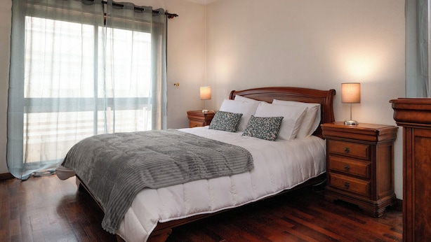 A neatly made bed with a grey blanket, two bedside tables with lamps, and a large window with sheer curtains. - Fort Beach Apartment (Figueira da Foz)