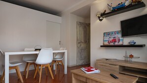 A modern living room with a white dining table, white chairs, a wooden coffee table, a TV, and decorative items on shelves. - Fort Beach Apartment (Figueira da Foz)