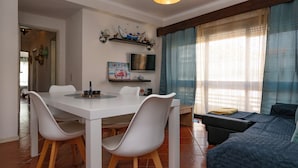 A dining area with a white table and chairs, a TV on the wall, and a sofa with a quilted cover. - Fort Beach Apartment (Figueira da Foz)